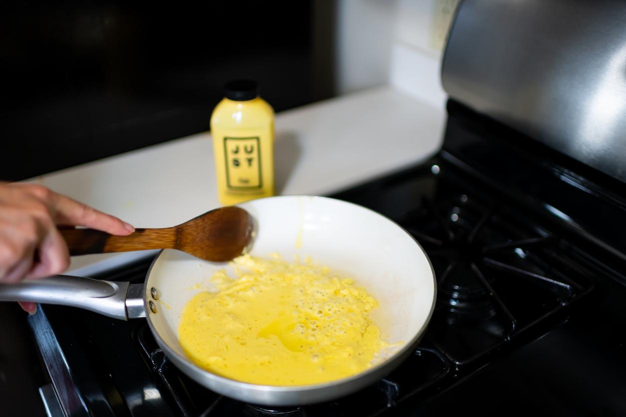 A person uses a wooden spoon to scramble the plant-based JUST egg substitute in a white ceramic pan atop a black stove.